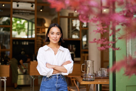 Portrait of young businesswoman in her own cafe, manager standing near entrance and inviting you, posing in white plain shirt and jeansの写真素材
