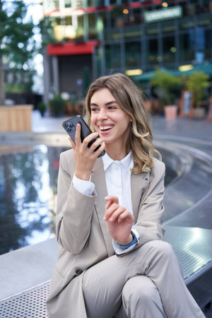 Smiling business woman record voice message, speaking into microphone on mobile phone, sitting near fountain on streetの写真素材