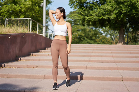Sportswoman jogging outdoors. Smiling asian fitness girl in wireless earphones, listening music and...