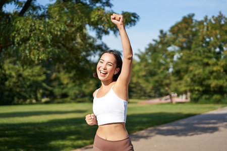 Hooray, victory. Smiling asian girl triumphing, celebrating achievement, running till finish, shouting from excitementの写真素材
