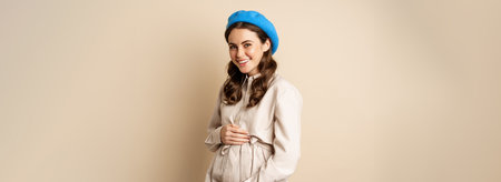Concept of pregnancy and surrogacy. Young gentle and tender woman with big belly. Smiling pregnant woman posing in trench coat against beige backgroundの写真素材