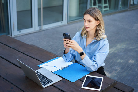Portrait of businesswoman sitting outdoors and working. Young corporate woman looking at her...