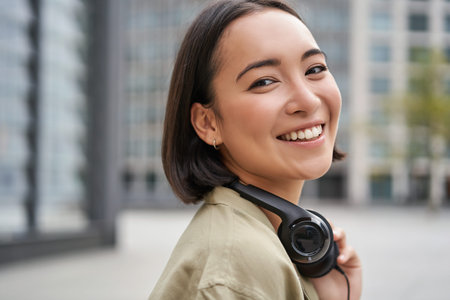 Portrait of happy asian woman in headphones, enjoys music while walking in city, smiling and...