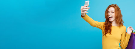 Shopping Concept - Close up Portrait young beautiful attractive redhair girl smiling looking at camera with white shopping bag and selfie. Blue Pastel Background. Copy spaceの写真素材