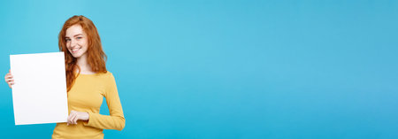 Close up Portrait young beautiful attractive ginger red hair girl smiling showing blank sign. Blue Pastel Background. Copy space.の写真素材