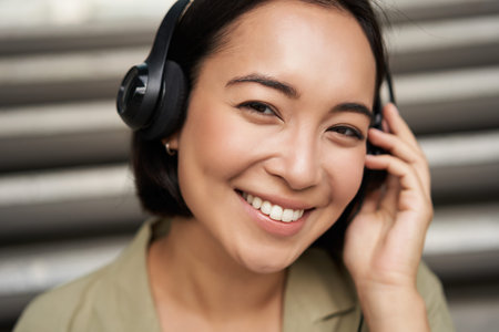 Close up portrait of smiling asian girl in headphones, listens to music outdoors, looking happy.
