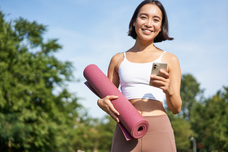 Portrait of young asian woman checking her phone during workout, walking in park with rubber mat, going to the gym, holding smartphoneの写真素材