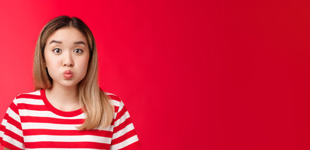 Close-up silly cute asian blond girl pouting making playful face, fold lips hold breath stand red background speechless, stay silent posing childish carefree look impressedの写真素材