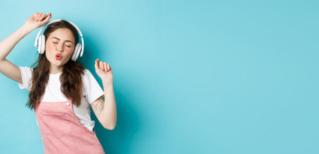 Beautiful young woman dancing and listening music in headphones, enjoying sound, standing over blue backgroundの写真素材