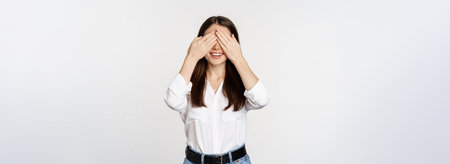 Smiling happy woman waiting for surprise, shut eyes with hands, standing blindsided against white...