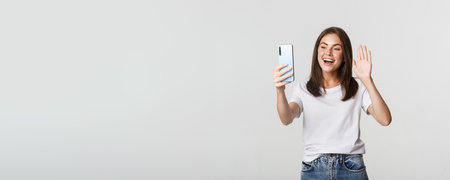 Friendly attractive girl saying hi, waving hand at smartphone during video call, having conversationの写真素材