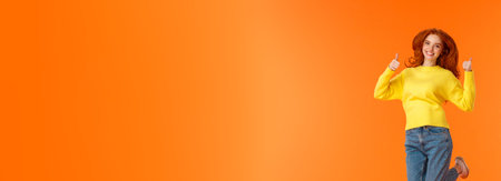 Shopping, fashion and women concept. Vertical full-length positive and cute redhead young modern hipster girl in denim jeans, winter yellow sweater jumping and showing thumbs-up in approval, smilingの写真素材