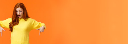 Fascinated and excited redhread hipster girl stare speechless with amazement and interest at bottom advertisement, look pointing down, drop jaw see cool product, shoppaholic found excellent saleの写真素材