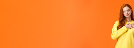 Tender and romantic, lovely young modern redhead woman 20s, touch heart and close eyes, remember dearest, touching moment, recall perfect date, keep memories in soul, orange backgroundの写真素材