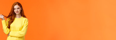 Gorgeous flirty and cute young modern redhead cheeky girl, rolling curl on hair and glancing camera curious, with interest and coquettish flirting smile, standing orange background intriguedの写真素材