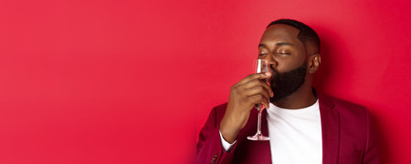Drunk african american guy kissing glass with champagne, celebrating new year, enjoying party, standing over red backgroundの写真素材