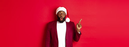 Christmas, party and holidays concept. Impressed Black man with beard, wearing santa hat, pointing finger left and gasping amazed, standing against red backgroundの写真素材