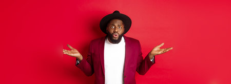 Confused and worried Black man spread hands sideways, staring at camera puzzled, asking what wrong, standing against red backgroundの写真素材
