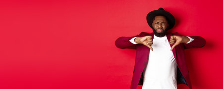 Disappointed Black man leave negative feedback, showing thumb down and grimacing from dislike and disgust, standing in party clothes over red backgroundの写真素材