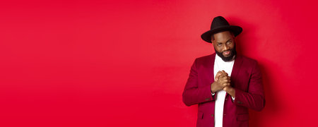 Cunning black man having an idea, smiling devious and rubbing hands, standing against red background...