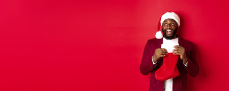 Cheerful Black man looking upper left corner and smiling, holding Christmas sock with gifts, standing over red backgroundの写真素材