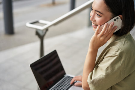 Portrait of asian girl, student sits on stairs with laptop, talks on mobile phone. Young woman makes a telephone call while working on computerの写真素材