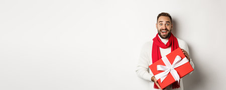 Concept of winter holidays. Surprised man receiving christmas gift and looking amazed, holding red wrapped box with present, standing amazed at white backgroundの写真素材