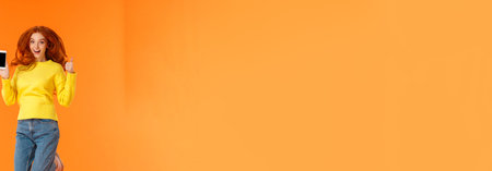 Advertising, technology and holidays concept. Vertical full-length shot cheerful gorgeous ginger girl, redhead woman jumping over orange background with smartphone, show thumb-upの写真素材