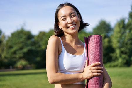 Portrait of asian girl smiles and laughs, stands with sports equipment, rubber mat, wears uniform for workout and exercises in parkの写真素材