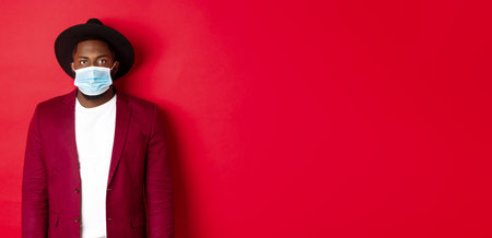 Covid-19 and fashion concept. Handsome black man in stylish hat, wearing face mask from coronavirus, looking serious, red backgroundの写真素材