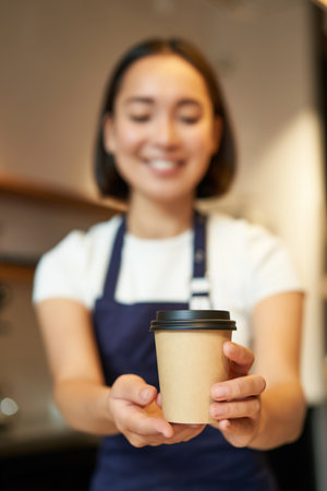 Smiling asian girl barista giving order to client, holding takeaway coffee cup, wearing apron, working behind counter in cafeの写真素材