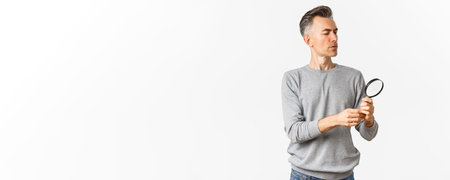 Portrait of thoughtful middle-aged man looking through magnifying glass, searching for something or reading, standing over white backgroundの写真素材