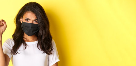 Concept of covid-19, social distancing and lifestyle. Close-up of fierce and confident african-american woman, being a protester in black lives matter movement, showing a fist, wearing face maskの写真素材