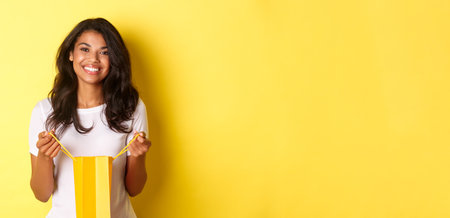 Portrait of happy and delighted african-american girl, open a shopping bad and smiling pleased, thanking for gift on holiday, standing over yellow backgroundの写真素材