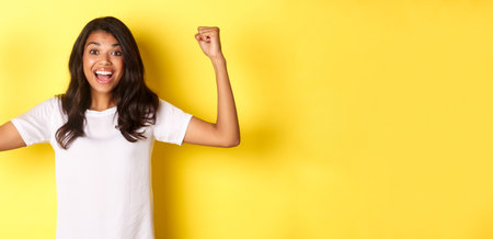 Image of happy african-american girl achieve goal and celebrating victory, raising hands up and smiling pleased, looking satisfied, standing over yellow backgroundの写真素材