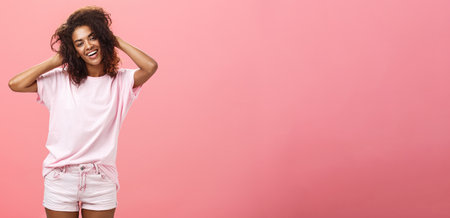 Girl enjoying nice weather chilling making photos for social network touching curly hair gently and carefree smiling broadly at camera standing over pink background happy and chillの写真素材