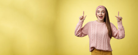 Ecstatic amused positive young 20s woman wearing casual sweater, headband dropping jaw amazed looking astonished, surprised pointing up excited, upbeat of seeing incredible promo over yellow wallの写真素材