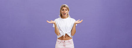 Perplexed and disappointment good-looking blonde female student in white t-shirt and pink shorts frowning shrugging with spread hands near shoulders saying what the hell over purple wallの写真素材