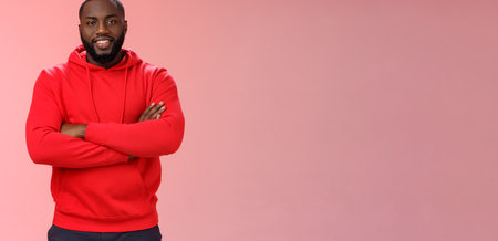 Attractive bearded african-american young guy wearing red hoodie cross arms chest smiling friendly showing friends new place grinning delighted buying own flat, standing proud pink backgroundの写真素材