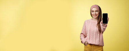 Best smartphone here you go. Charming outgoing young pretty woman in knitted sweater, headband extending hand with phone showing gadget screen smiling recommending cellphone over yellow wallの写真素材