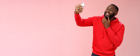 Cheeky cool african american confident bearded guy in red hoodie taking selfie update date app profile photo holding smartphone raised touching face sassy smiling look phone display flirtyの写真素材