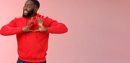 Someone love ya. Portrait enthusiastic creative cute black boyfriend wearing red hoodie show heart sign smiling broadly confessing love sympathy look passionate, express romance pink backgroundの写真素材