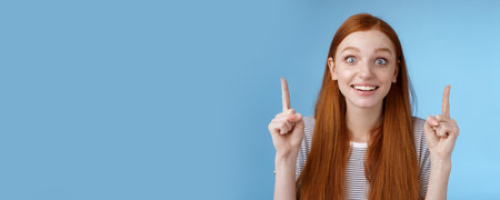 Thrilled happy young rejoicing redhead girl get awesome chance look surprised astonished smiling broadly wide eyes impressed incredible offer pointing up index fingers excitement, blue backgroundの写真素材