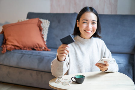 Smiling cute asian woman using credit card and smartphone, paying bills online, holding mobile...