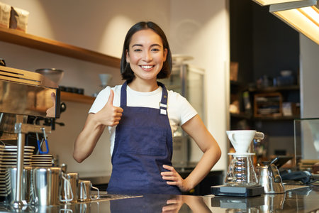 Smiling asian barista, girl in cafe uniform, shows okay sign, recommends their coffee, approve new flavours, standing in apronの写真素材