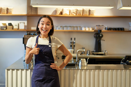 Smiling girl student working part-time in cafe, barista shows thumbs up, wears apron, stands near coffee shop counterの写真素材