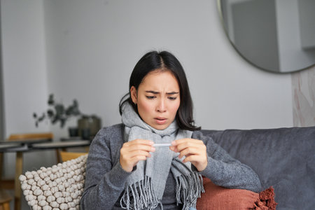 Shocked woman looks concerned at her thermometer, catching cold, has fever, sitting on sick leave at homeの写真素材