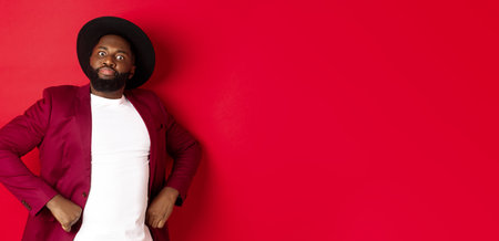 Funny african american man trying to act serious, holding hands on waist and staring at camera scary, standing over red backgroundの写真素材