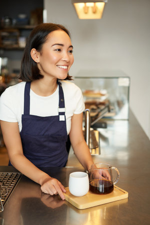 Vertical shot of smiling girl barista serving coffee, making batch brew, filter for client in cafe, wearing blue apron behind counterの写真素材
