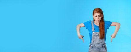 Moody upset cute redhead woman feel uneasy, frowning look pointing up jealousy and regret, cannot afford awesome new product, express sadness and sorrow, stand blue background gloomyの写真素材